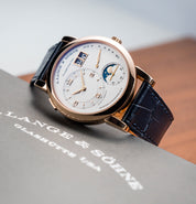 Lange 1 with dark navy blue alligator strap, gold casing, white dial, subdials, and moon phase rests on an A. Lange & Söhne book.