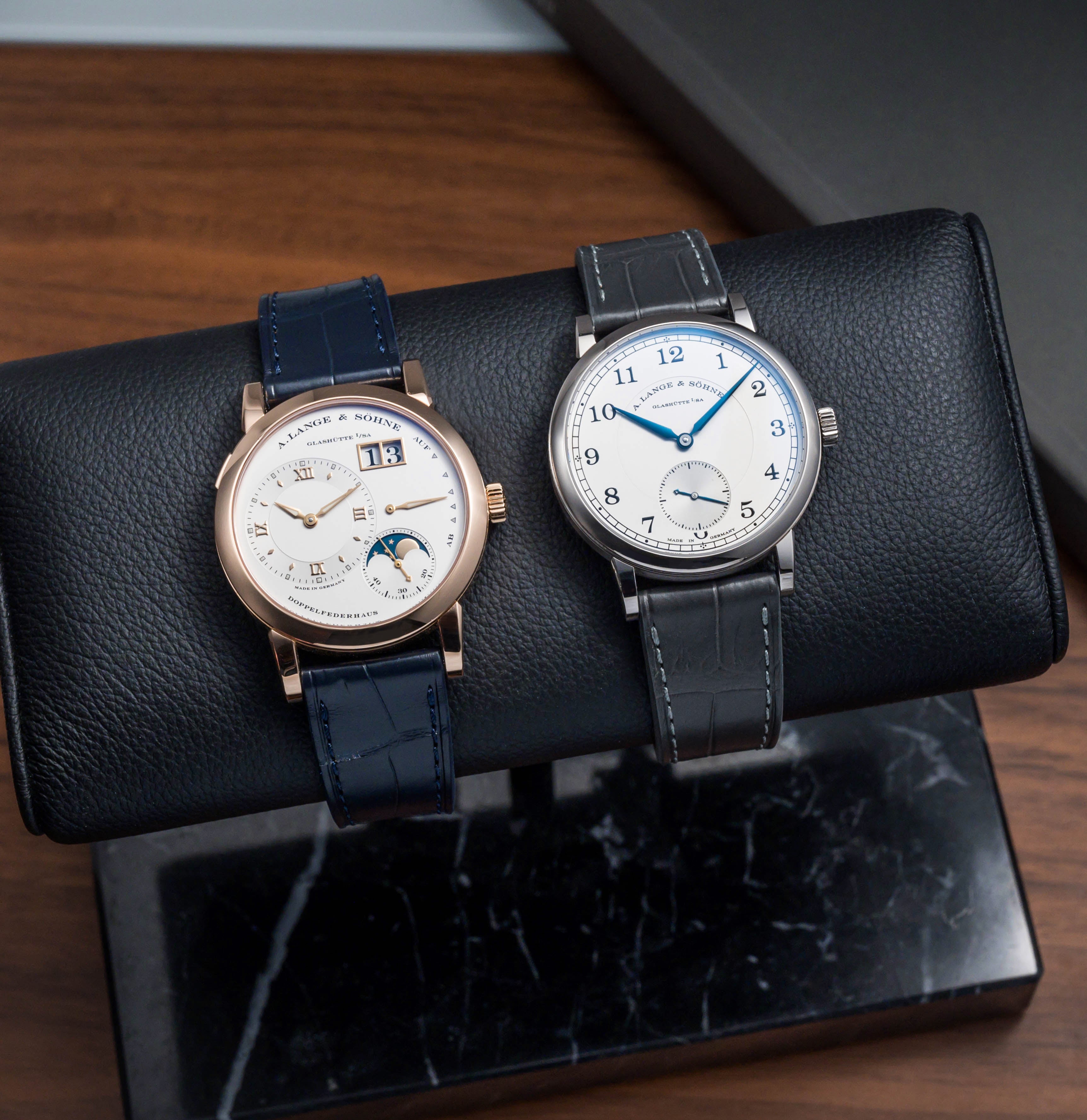 Classic Lange 1 and 1815 rest on a black cushion. Lange 1 pairs with RSM Navy Alligator Strap, 1815 with gray leather, both offering classic dials.