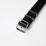 Close-up of the Herringbone Twill in Artillery Black NATO watch band by RSM Watch Strap, with a silver buckle and two loops on a white background.