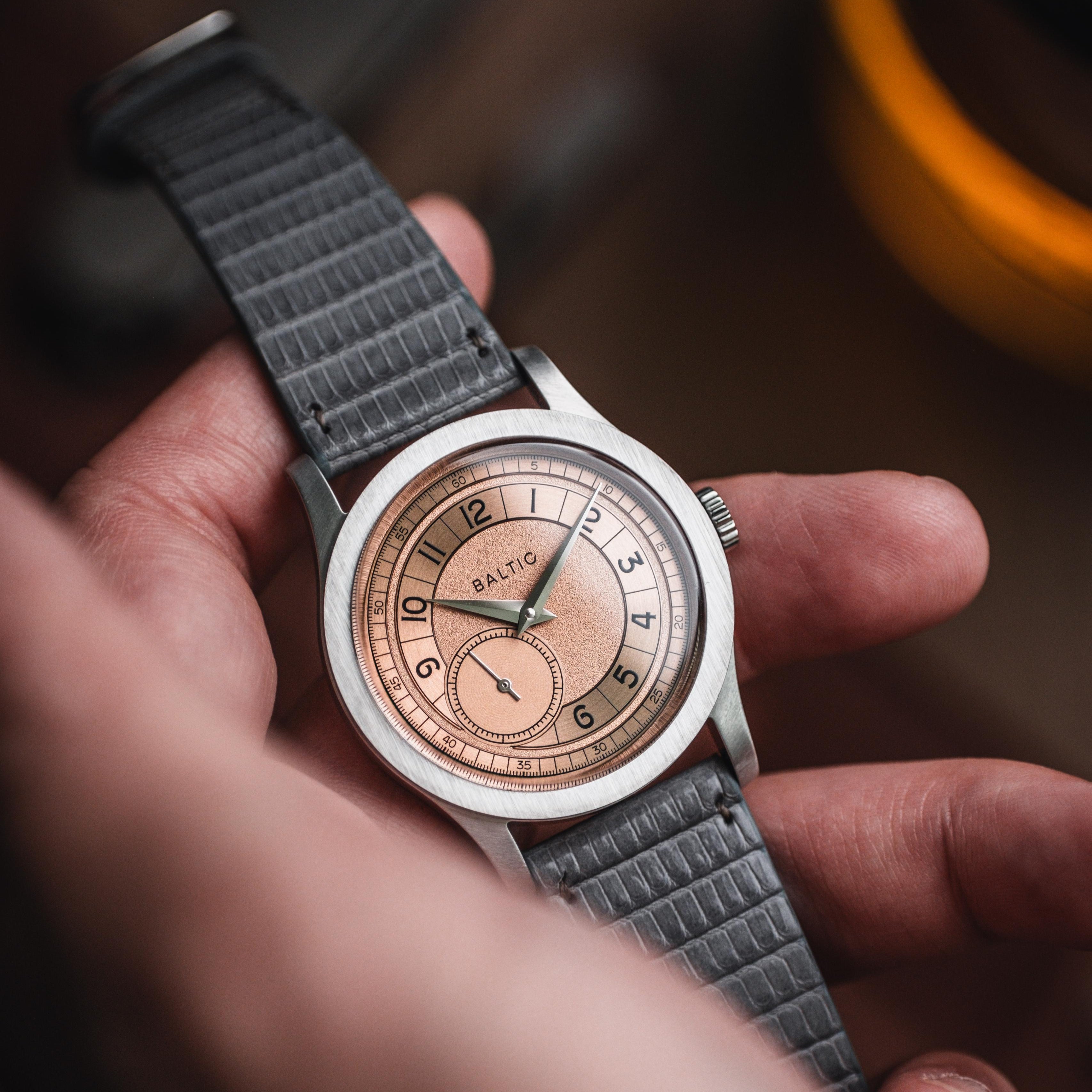 A person holds a timepiece with grey RSM Spanish lizard leather strap, copper-toned Baltic dial, black numerals, and hands, blurred background.