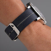 A wrist flaunts the RSM Engraved Stripe in Blue Grey Patina, featuring a dark blue strap, cream stitching, glossy buckle, and metallic case on gray.