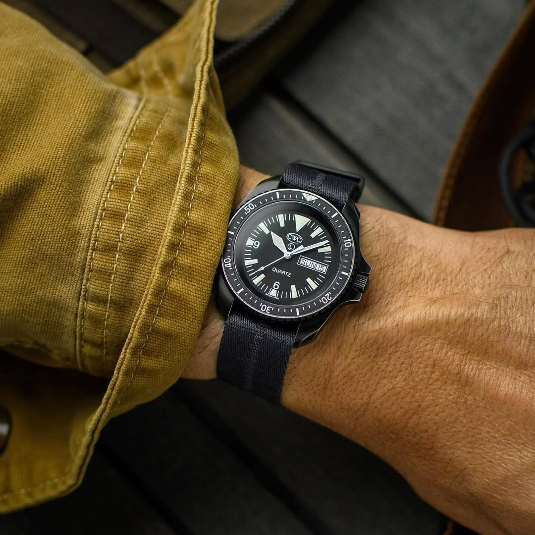 The Stealth Black Engraved Stripe RSM Strap with a black dial rests on a wrist in a brown jacket, with a jacquard weave bag in the background.