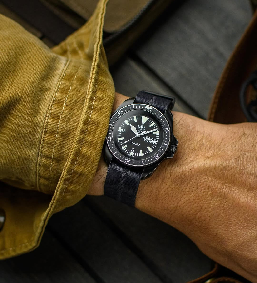 Black CWC dive watch is worn with RSM Watch Strap's engraved stripe's stealth black color with center line in black also. Wrist shot has mustard canvas sleeve with timber background.