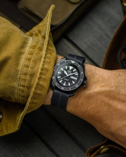 Black CWC dive watch is worn with RSM Watch Strap's engraved stripe's stealth black color with center line in black also. Wrist shot has mustard canvas sleeve with timber background.