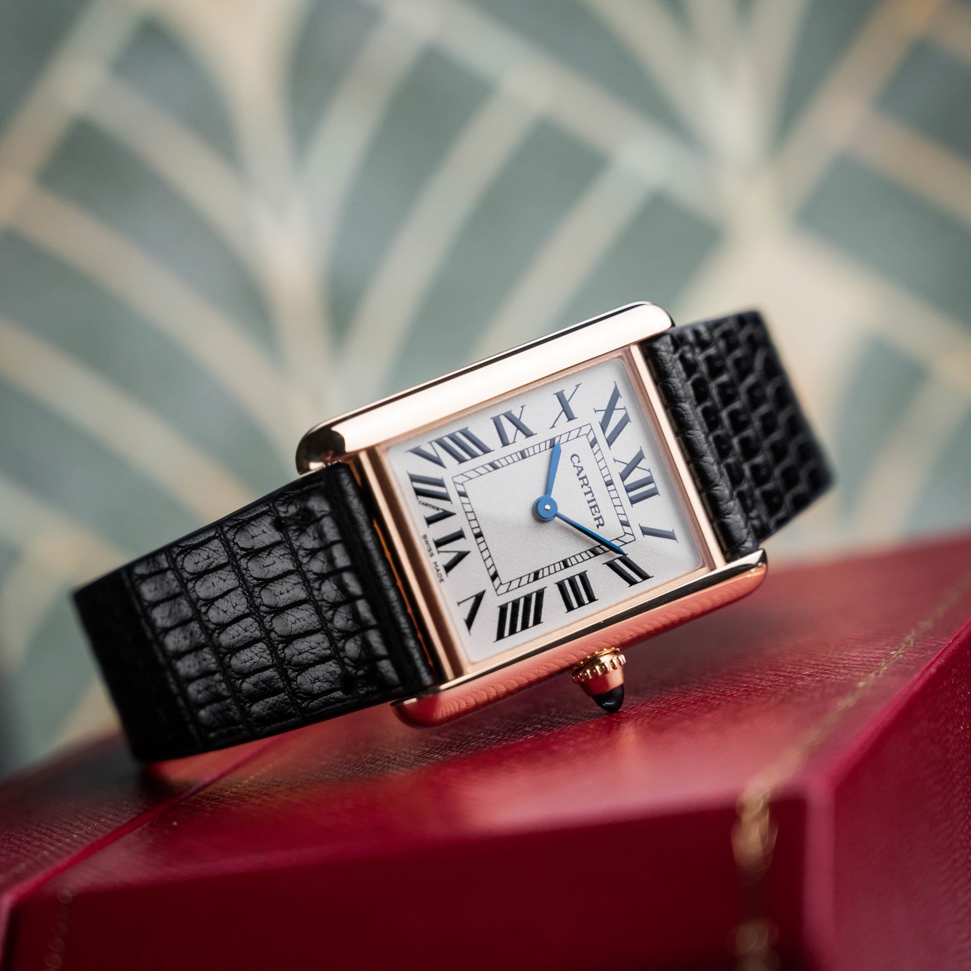 Cartier Tank gold case on RSM's black Spanish lizard strap with minimal stitching, resting on a Cartier red box with an art deco leaf pattern.