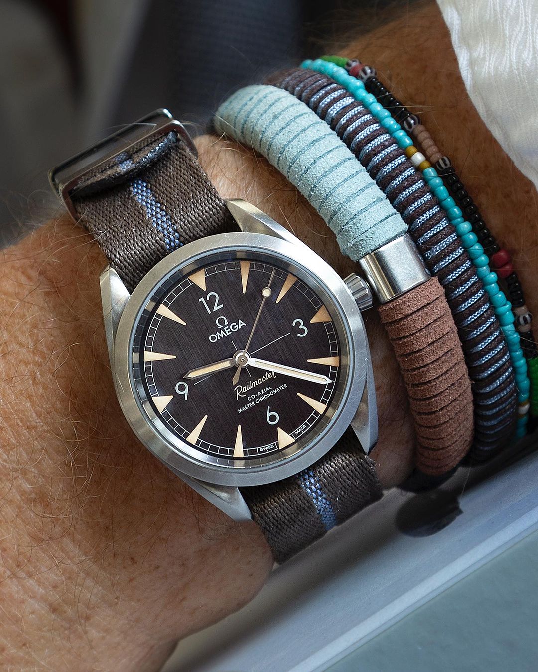 A man's wrist with an Omega watch, brown dial, white numerals, and RSM Engraved Stripe in Blue Coffee, paired with beaded bracelets.