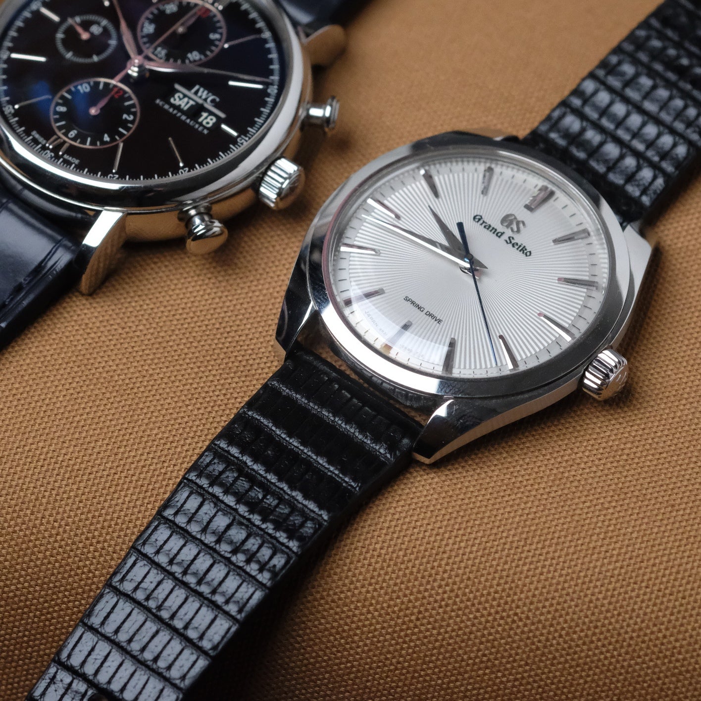 Two elegant timepieces on brown fabric: Grand Seiko SBGY003 with black Spanish leather, and IWC Portofino with Dark Navy Alligator strap.