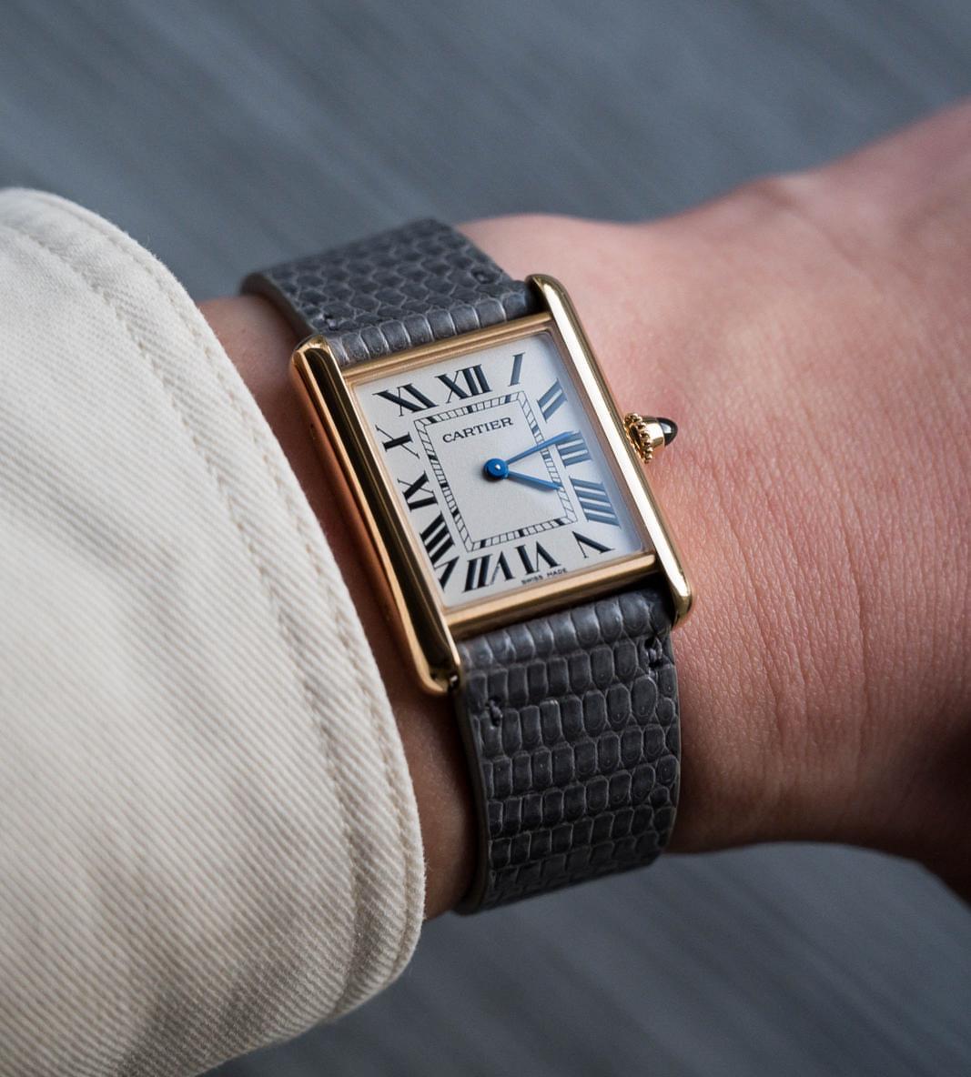 Wrist shot of Cartier Tank gold case, white dial, black Roman markers, blue steel hands, worn with lizard strap by RSM.