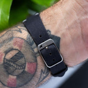 Close-up of the black coal Fine Canvas watch strap walrus logo buckle on a tattooed wrist, featuring a sleek 316L stainless steel buckle for durability and style.