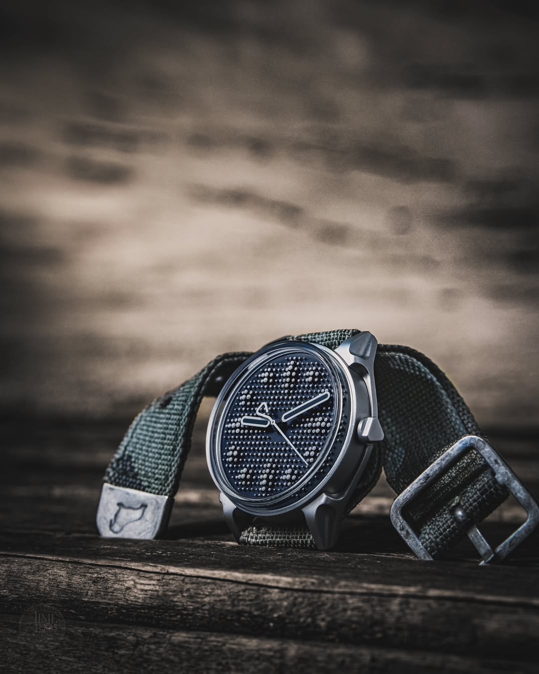 Micro brand watch Kollokium 01 placed on a timber table with matte steel case and its dial with multiple pin heads forming hour markers. The watch is fitted on the RSM camo strap with distressed steel hardware.