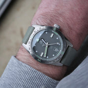 A wrist displays the Ollech & Wajs M-110 with RSM Fine Canvas Safari Green watch strap, silver markers, date at 3 o'clock, rotating bezel, and quick-release spring bars.