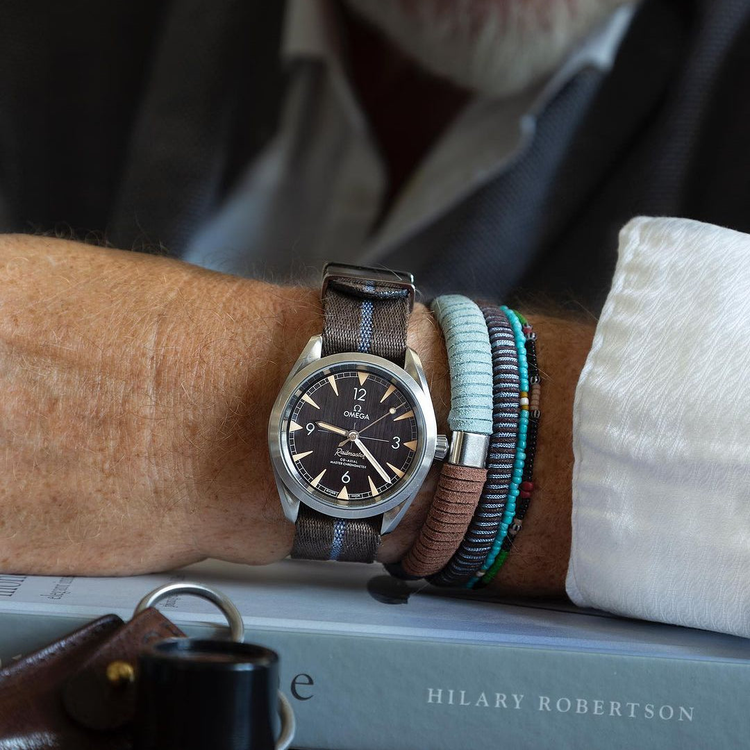 A wrist with an Omega watch and RSM's Engraved Stripe in Blue Coffee strap, surrounded by bracelets and a Hilary Robertson book