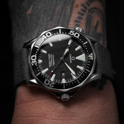 A tattooed hand showcases the Omega Seamaster Peter Blake Diver 300m 2254.50.00 with a wave pattern, luminescent markers, and an RSM Fine Canvas watch strap.