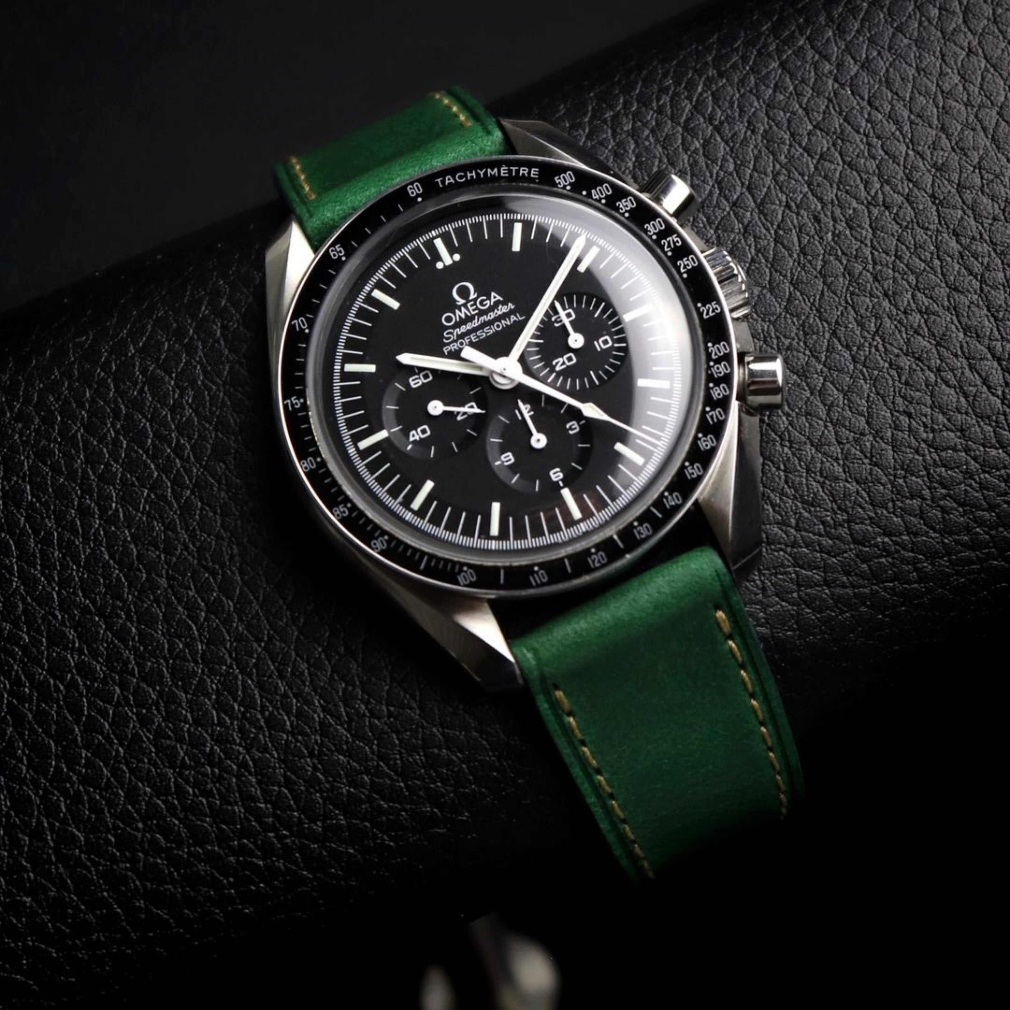 Omega Speedmaster with RSM's Green Buttero strap featuring beige stitching rests on a black leather watch stand, showcasing its soft, supple texture.