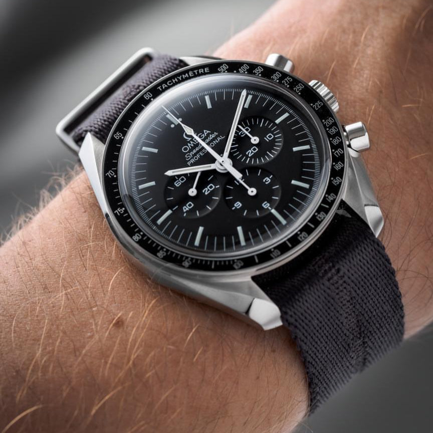 A wrist with an Omega Speedmaster featuring a black jacquard weave dial, subdials, and tachymeter bezel, paired with RSM Stealth Gray strap.