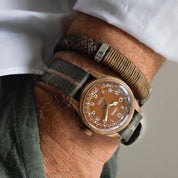 Oris Big Crown bronze watch wrist shot with brown leather and coiled nylon strings. RSM's engraved stripe in green, beige, and orange matches the outfit.