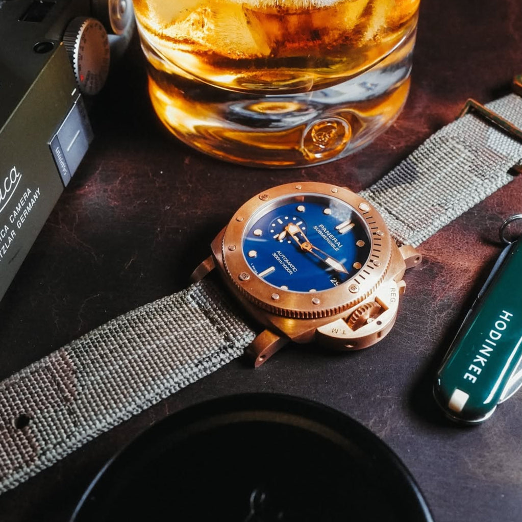 Blue dial Panerai Bronze on a 22mm RSM camo 2-piece strap with distressed bronze hardware. The camo weaving complements the watch's military style.