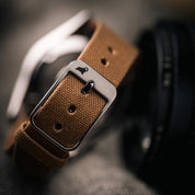 Close-up of RSM's Roasted Tea Fine Canvas Strap, showcasing tight Japanese canvas grains and 316L Stainless Steel buckle, with a bokeh background.