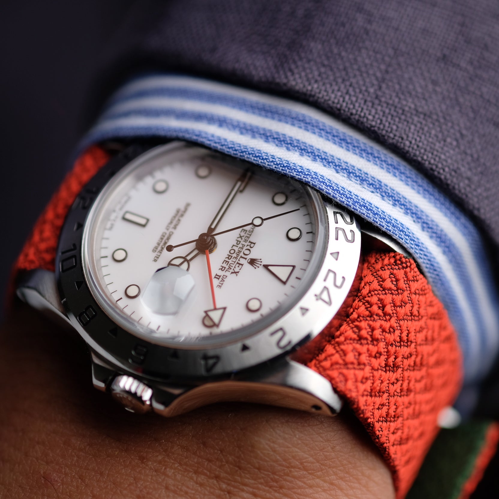 Close-up of an RSM watch with a white dial, black numerals, and an orange 2-Piece Silk Strap. Silver hands, indices, and a gray sleeve with a striped cuff.