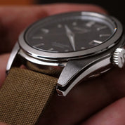 A close-up wristwatch with a Fine Canvas Strap in Pine Khaki by RSM Watch Strap shows a stainless steel case, dark dial, and polished bezel resting on a hand.