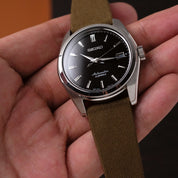 A Seiko wristwatch with a black dial, silver casing, and RSM Pine Khaki Fine Canvas Strap made from Japanese cotton yarns is held in a hand.