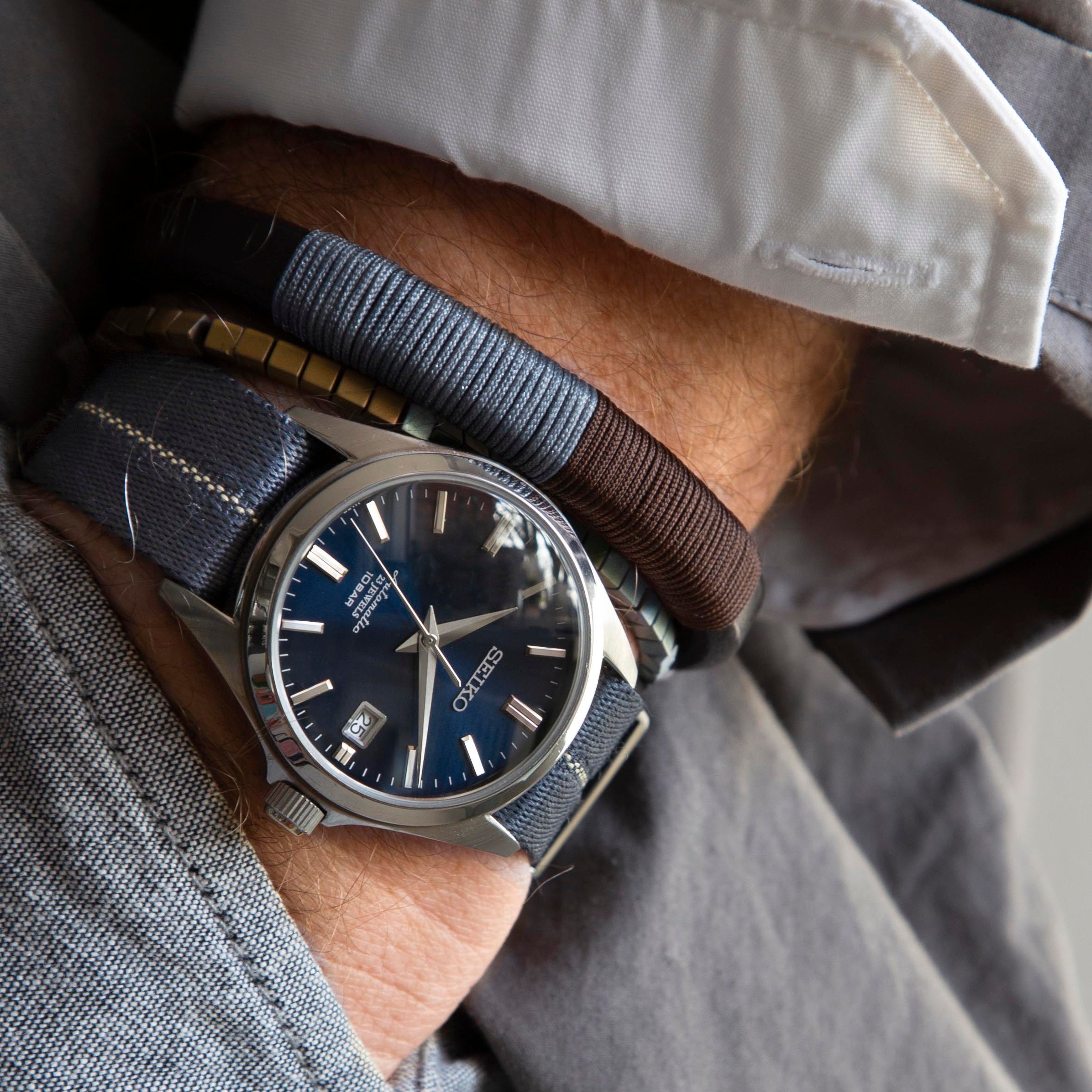 An individual sports a dark blue Engraved Stripe in Blue Grey Patina wristwatch with a date feature, complemented by RSM fabric strap and bracelets.