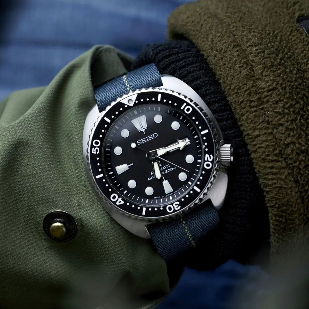 A wrist adorned with a Seiko diver’s watch, black dial, and RSM Engraved Stripe in Blue Grey Patina strap, paired with a green jacket and black glove.