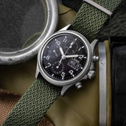 Close-up of a wristwatch with a black dial, white markings, chronograph subdials, and Herringbone Twill in Olive Drab RSM Watch Strap.