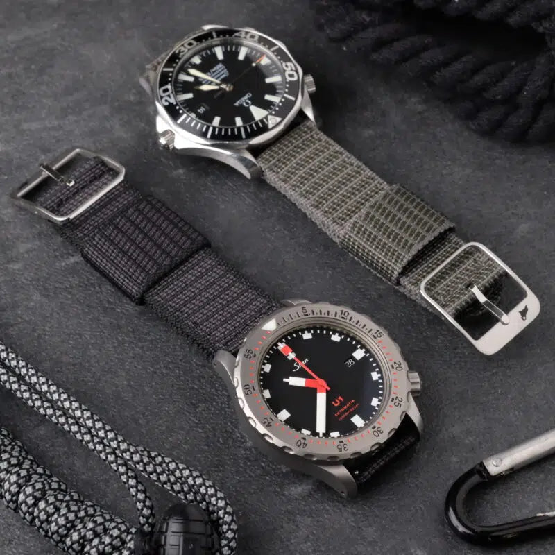 Two stylish wristwatches on a dark surface: left with RSMs X-Hatch (Black) strap, right with gray strap, black dial, red accents, ropes, and carabiner.