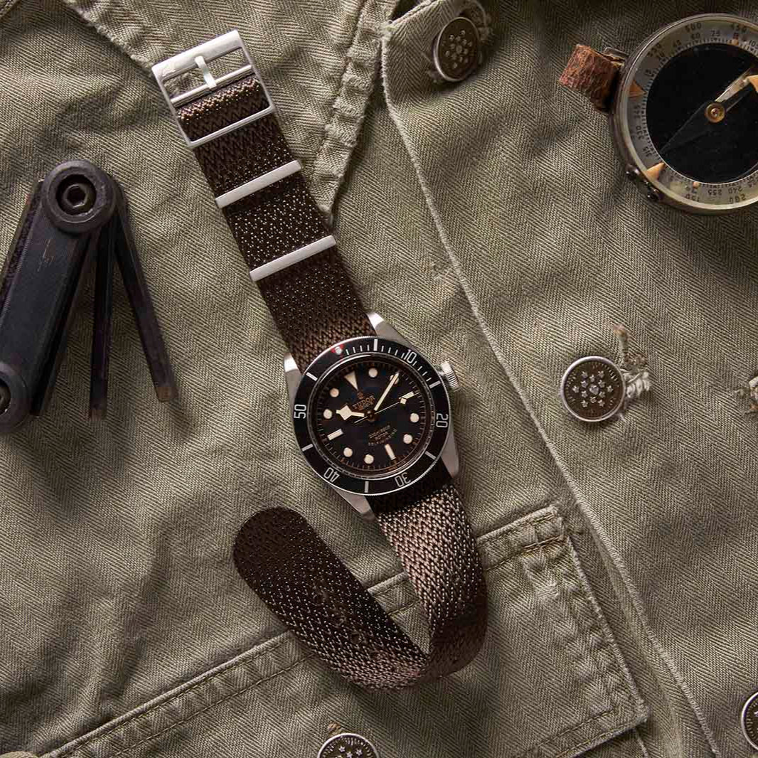 RSM Herringbone Twill Tobacco Brown strap rests on a green military jacket with a compass and multi-tool, evoking an adventurous vibe.