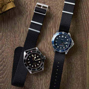 Two Herringbone Twill watches with black NATO bands rest on wood; one has a vintage black dial, the other a modern blue dial.