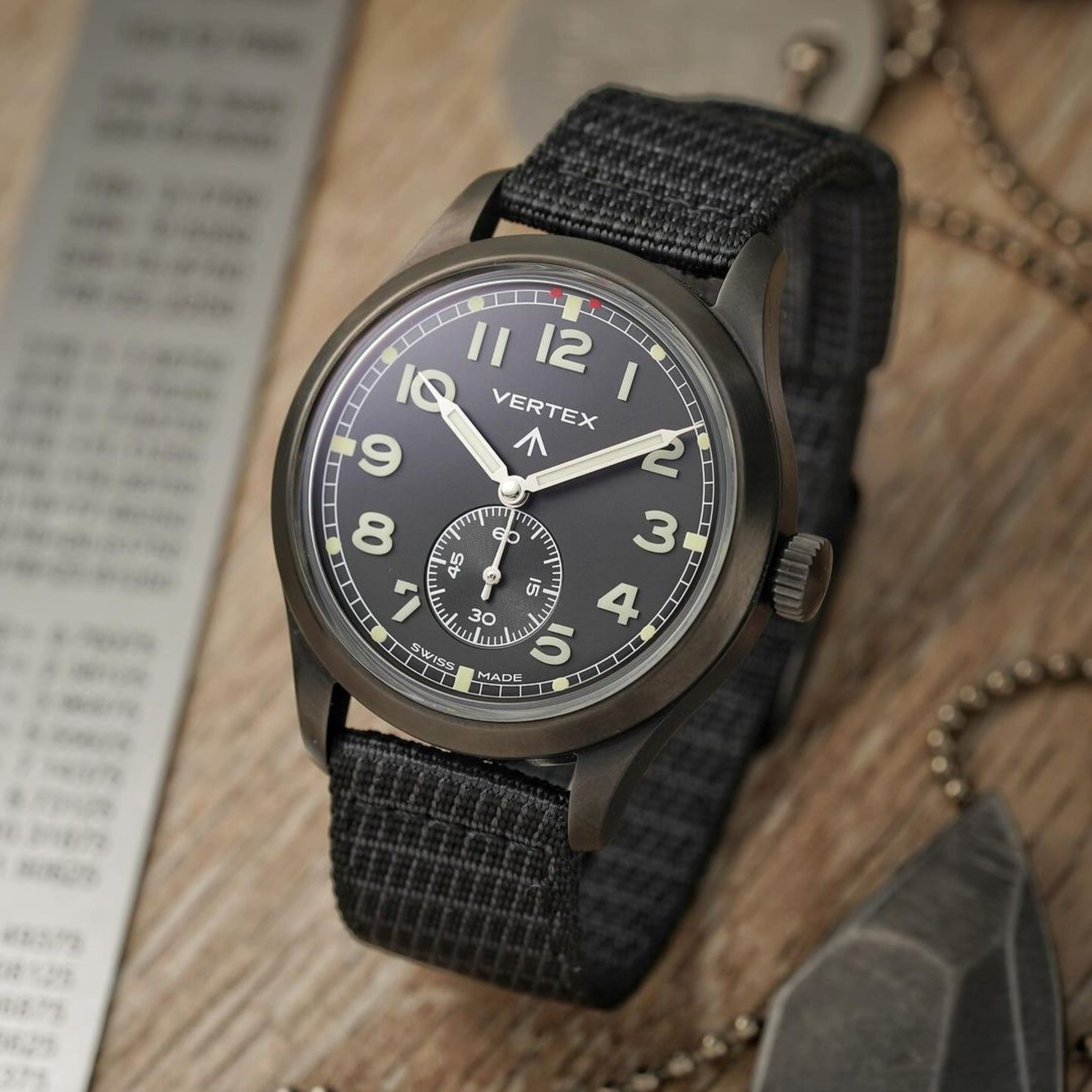 A military-inspired wristwatch with a black dial and X-Hatch Watch Strap from RSM is displayed on wood with a ruler, tag, and chain nearby.